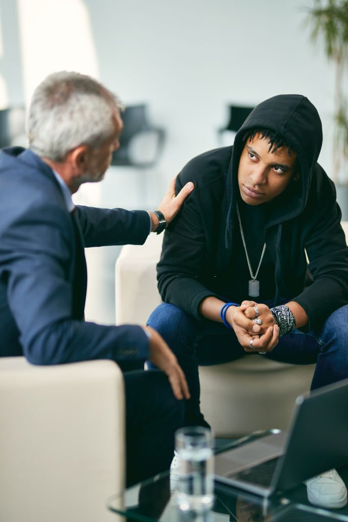man with grey hair wearing gray suit putting his hand on a young mans, wearing a black hoodie, shoulder