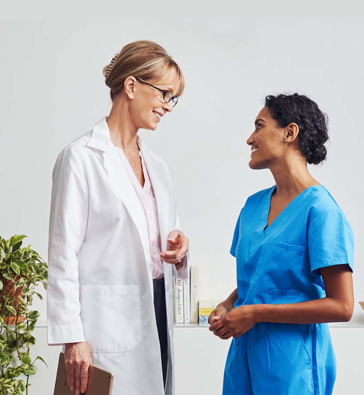 tall blonde woman in lab coat smiling and looking at woman in blue scrubs and dark brown hair