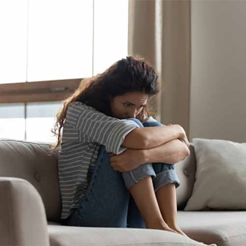 woman with long black hair sitting on gray couch hugging her knees while looking out window by Allied Healers